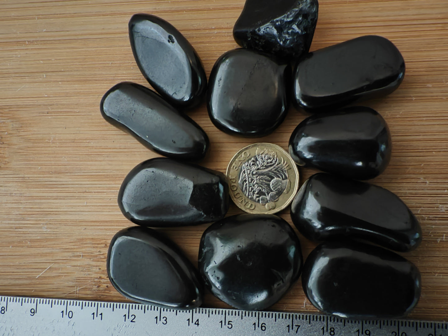 Ten smooth, polished shungite stones arranged on a light wood surface, with a coin in the center for scale and a ruler visible along the left edge.
