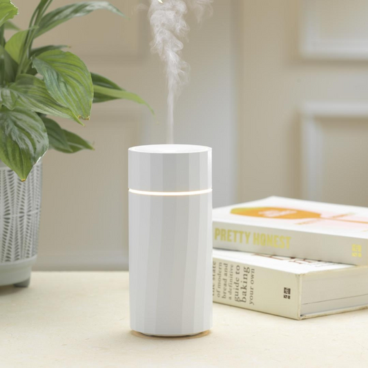 A white cylindrical aroma diffuser from MadeByZen NOMAD, emitting a mist, placed on a table near a plant and a book.