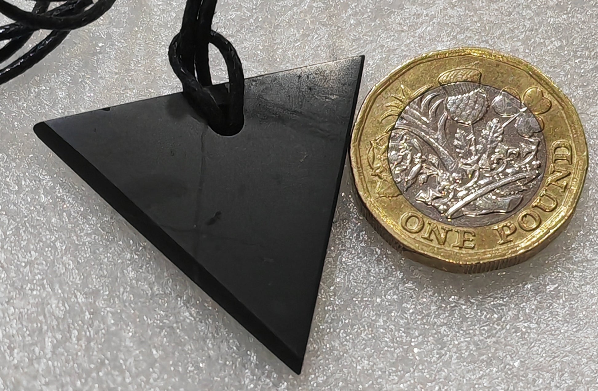 A triangular black shungite stone pendant on a black cord, shown next to a UK two-pound coin for scale on a textured white background.