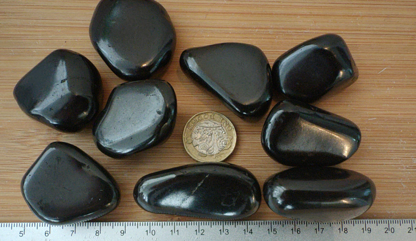 Nine smooth, polished shungite stones arranged on a light wood surface next to a metal ruler and a coin for scale.