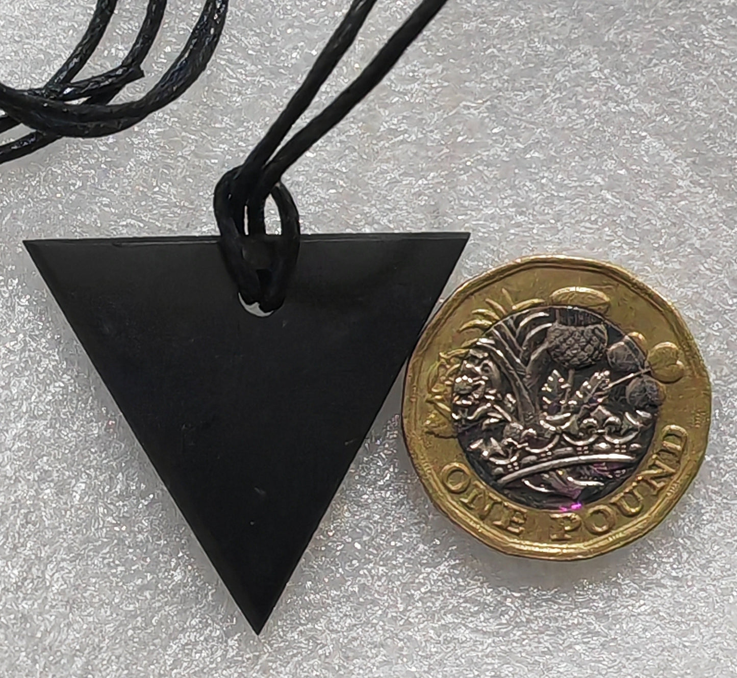 A black shungite triangular pendant necklace on a black cord, shown next to a British two-penny coin for scale on a textured white background.