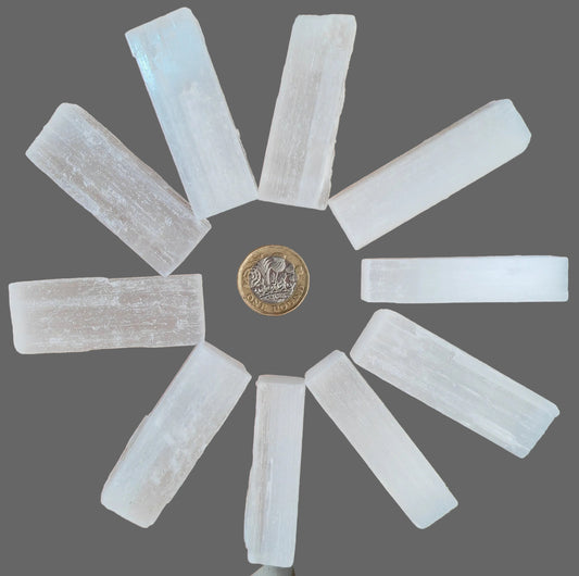 Nine translucent white selenite crystal sticks arranged in a circular pattern on a wooden surface, with a British coin placed in the center for scale.