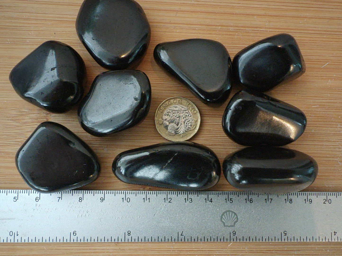 Nine smooth, polished shungite stones arranged on a light wood surface next to a metal ruler and a coin for scale.