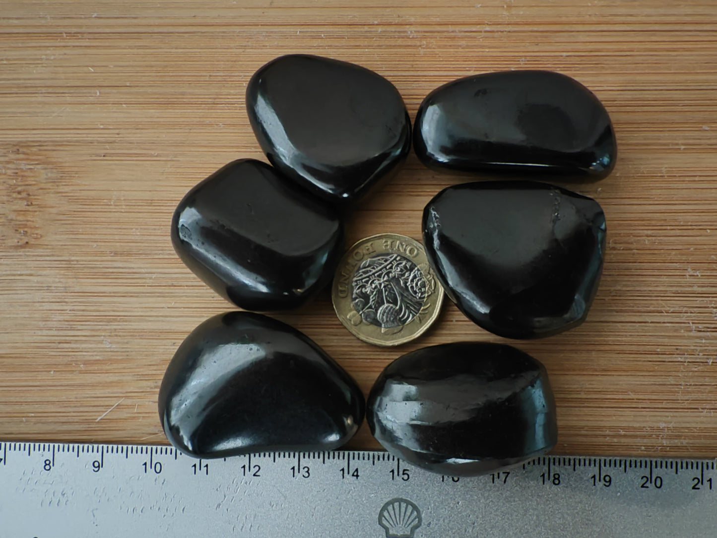 Six smooth shungitepolished stones arranged in a circle around a British coin on a light wood surface, with a metal ruler visible along the left edge for scale.